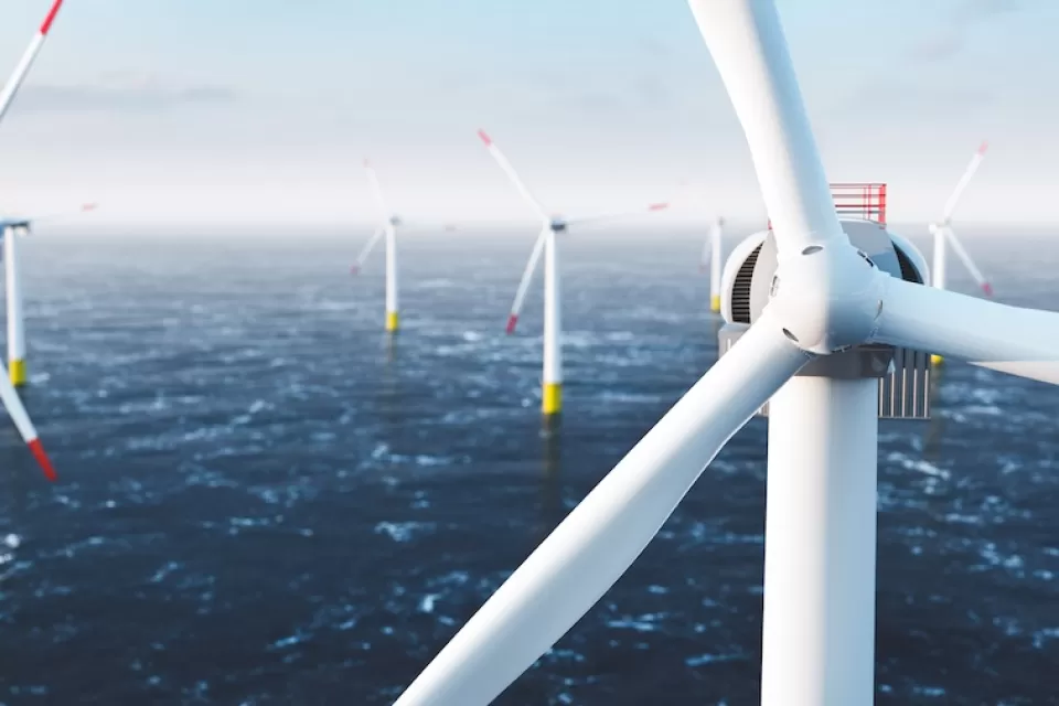 Close-up of top of white offshore wind turbine with other turbines in sea in background.